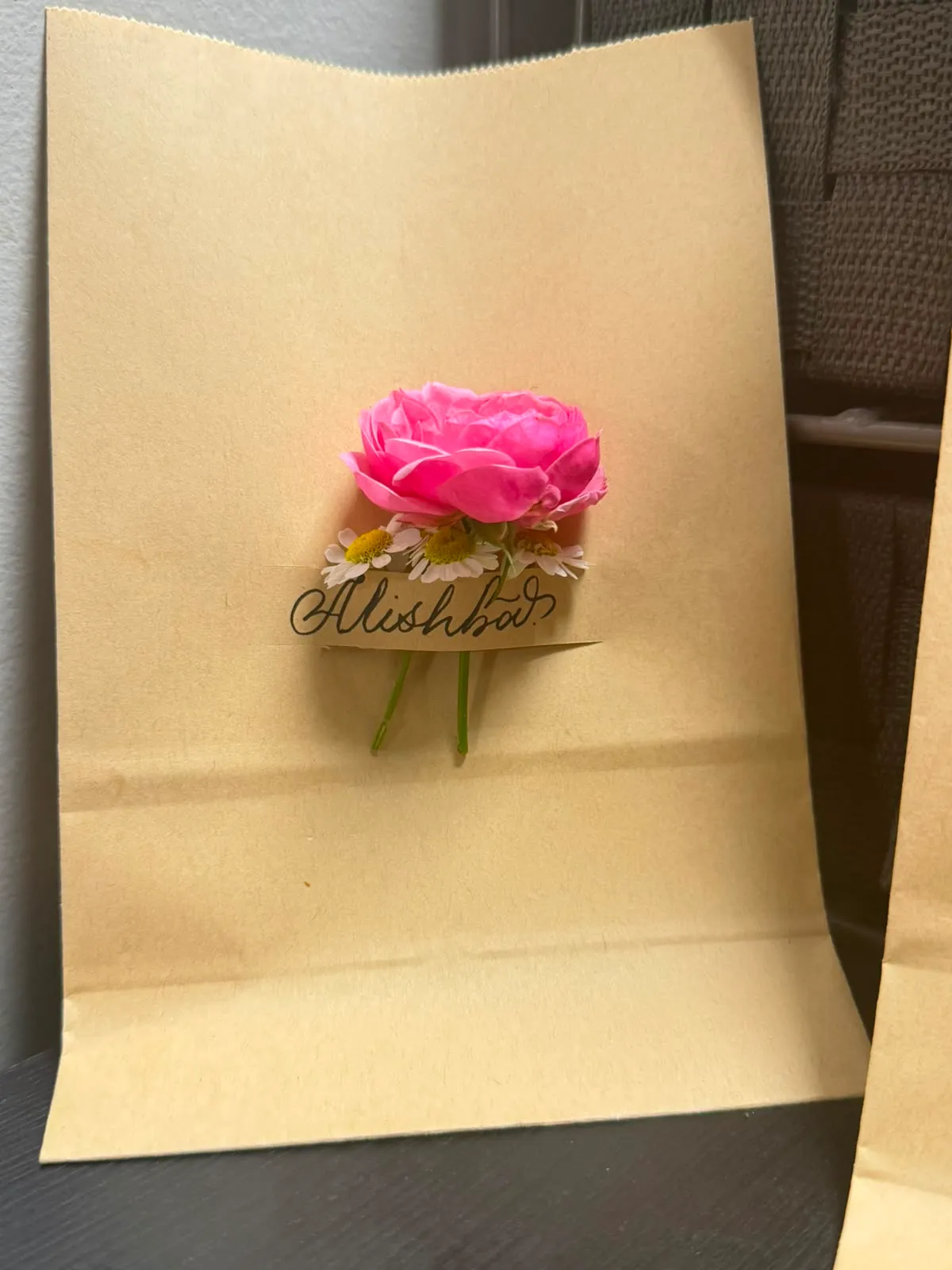 Kraft gift bag lettered “Alishba” with a pink rose and daisies