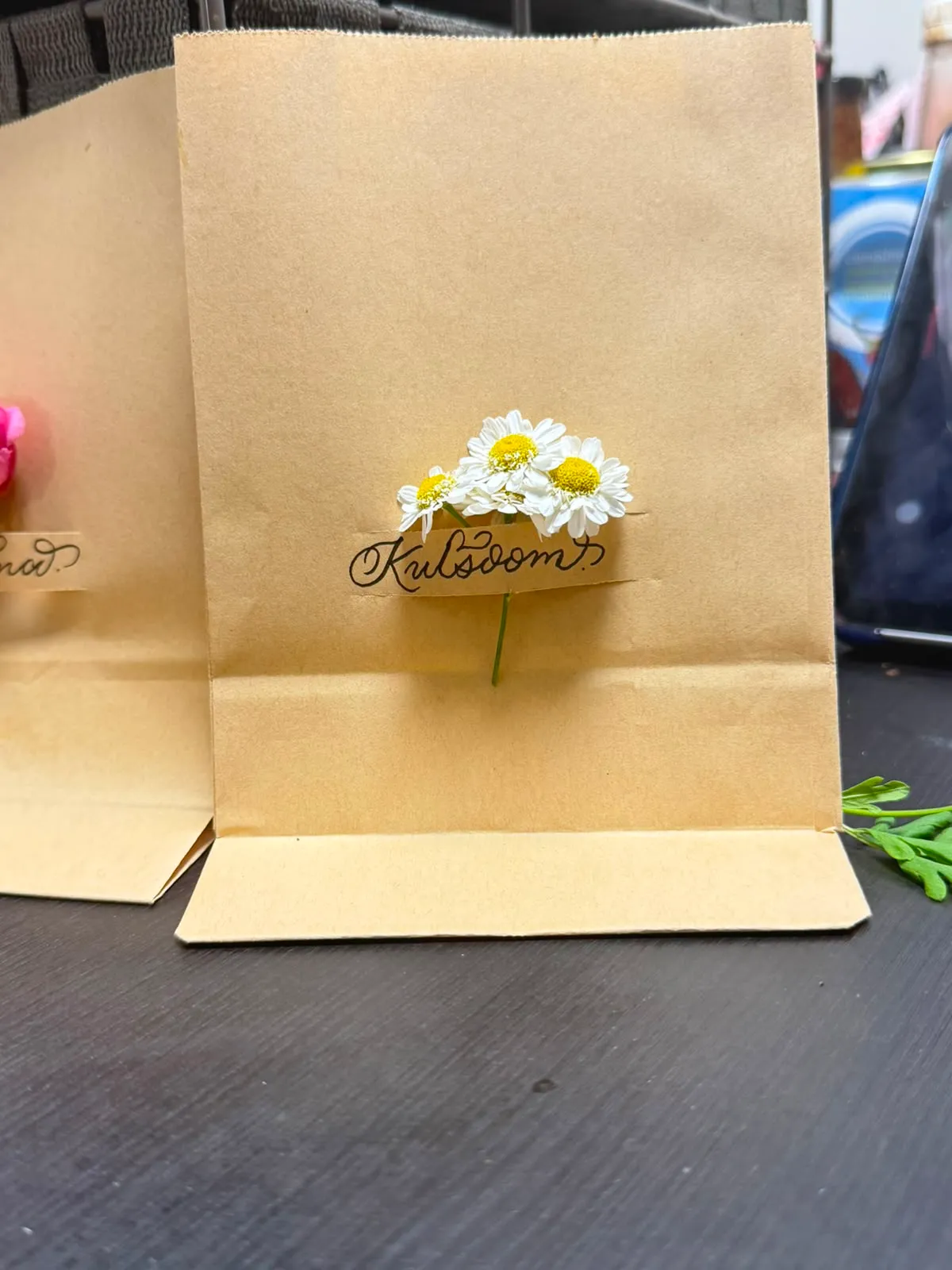 Kraft gift bag lettered “Kulsoom” with fresh daisies